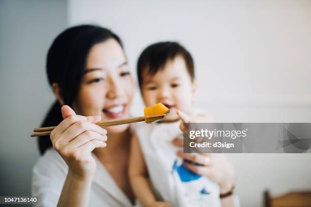 smiling mother eating pumpkin with chopsticks while curious baby girl pointing at it - asian family cafe stockfoto's en -beelden