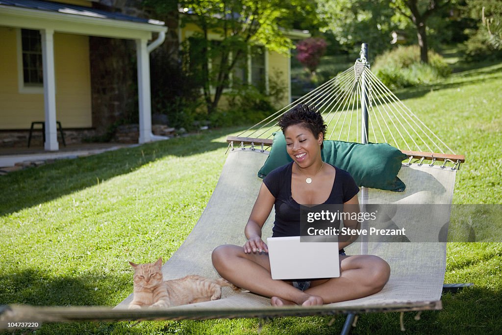 Black woman sitting in hammock with laptop and cat