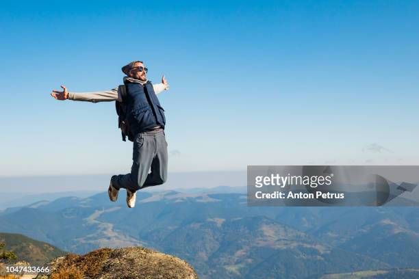 young man jumping on top of a mountain against the sky - hipster brille stock-fotos und bilder