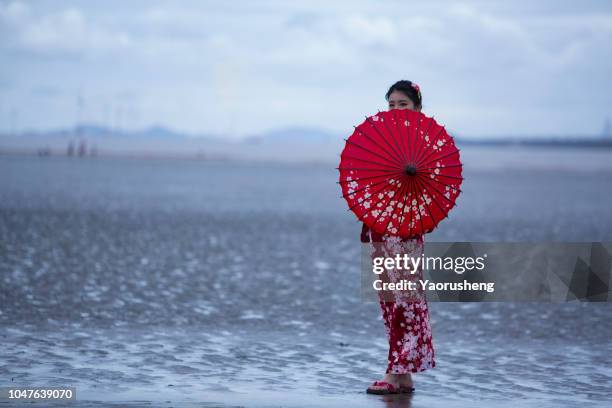 portrait of young asian woman wearing kimono with red umbrella,walking on beach - costume designs stock pictures, royalty-free photos & images