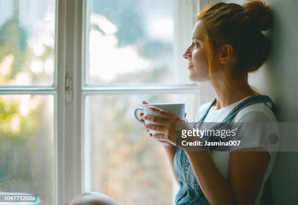 hermosa mujer joven mirando por la ventana y tomando café en la mañana - tranquilidad fotografías e imágenes de stock