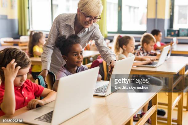 gelukkig rijpe leraar schoolkinderen bijstaan bij het gebruik van computers op een klasse. - elementary school classroom stockfoto's en -beelden