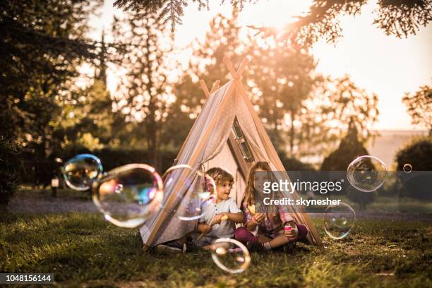 niños pequeños jugando con la varita de la burbuja frente a una tienda de campaña al aire libre. - bubbles happy fotografías e imágenes de stock