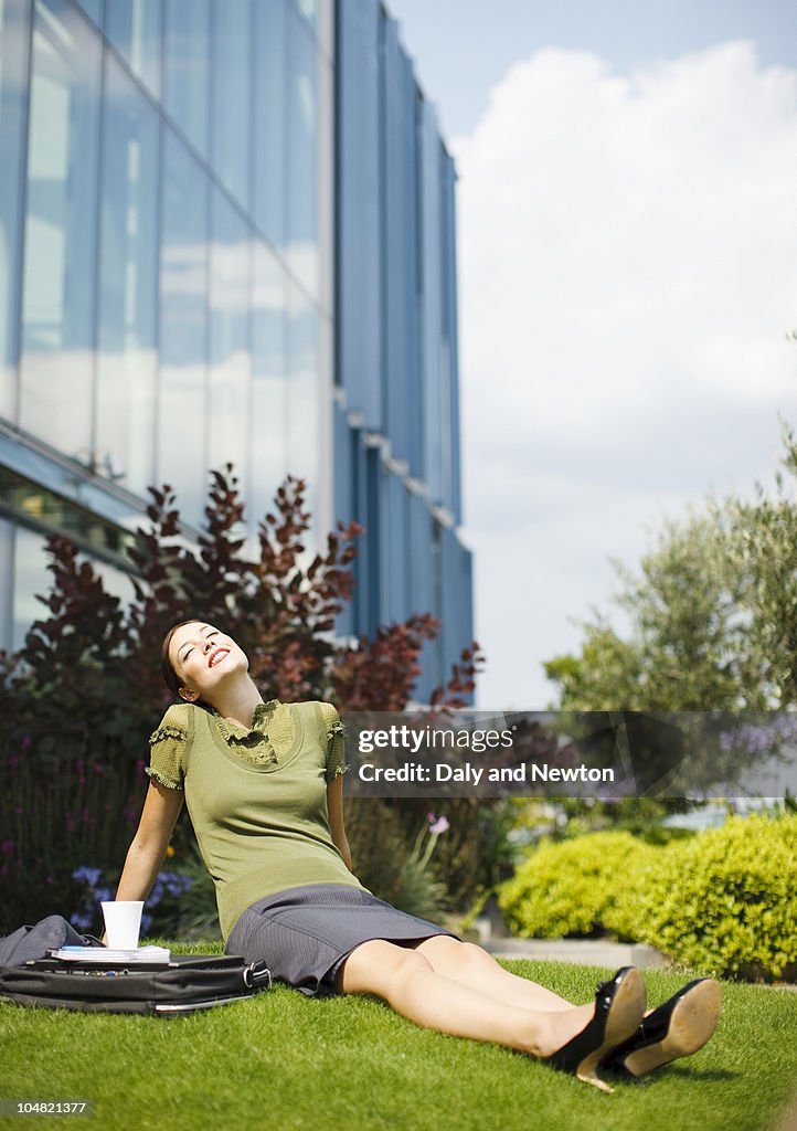 Smiling businesswoman sitting in grass and basking in sunlight outside office building