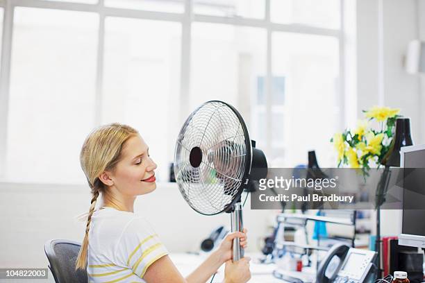 femme souriante, assis en face de fan dans le bureau - ventilateur électrique photos et images de collection