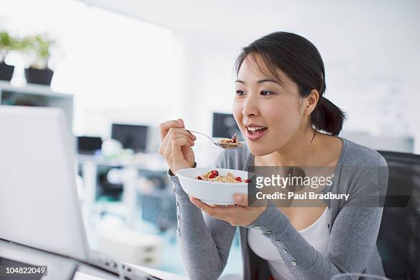 businesswoman eating cereal and looking at laptop in office - breakfast work stock pictures, royalty-free photos & images