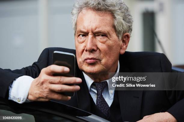 portrait of a senior man standing beside his car checking on his smartphone - grumpy old man stock pictures, royalty-free photos & images