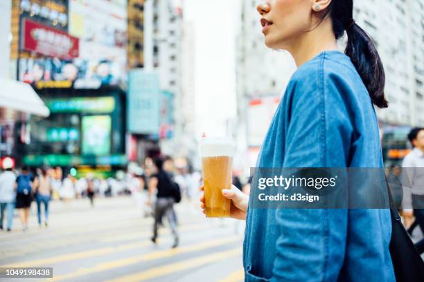 close up of woman holding a bottle of iced drink and walking in busy downtown city street against pedestrians and city skyscrapers - taiwanese ethnicity stock-fotos und bilder