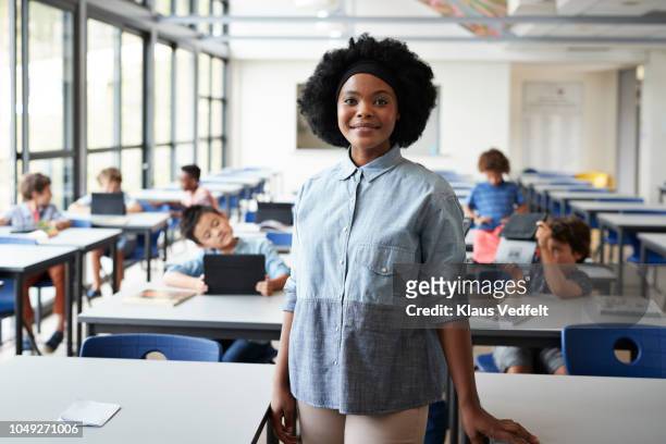 portrait of female teacher standing in classroom with students in background - teacher trigger stock pictures, royalty-free photos & images