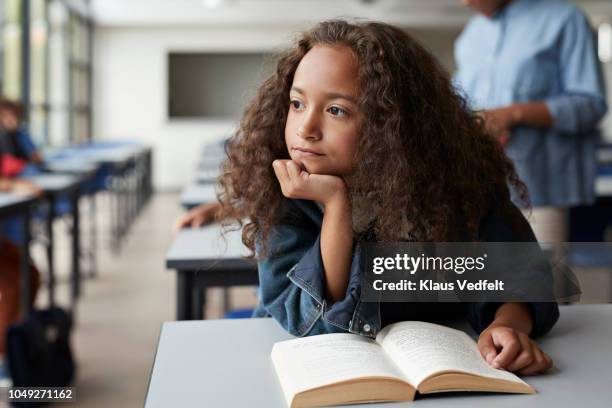 girl sitting with book and looking thoughtful out of window - unhappy school child stock pictures, royalty-free photos & images