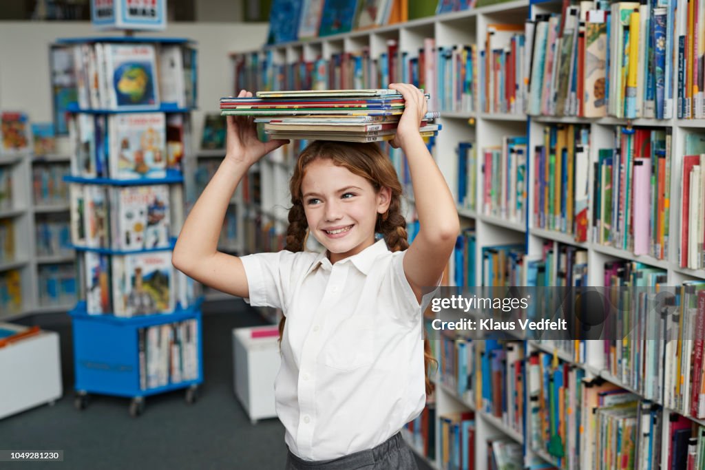 Cute schoolgirl smiling & balancing stack of books on the head at library