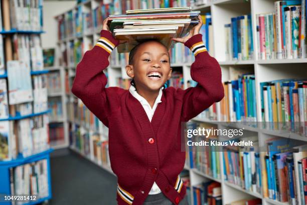 cute schoolgirl smiling & balancing stack of books on the head at library - africa child stock-fotos und bilder