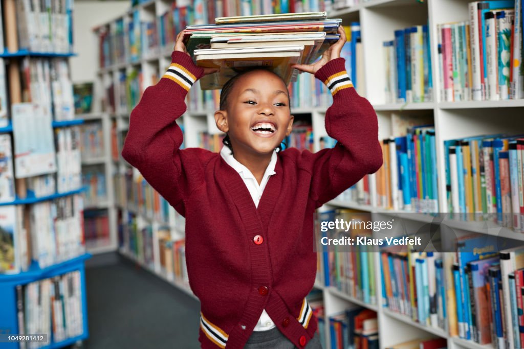 Cute schoolgirl smiling & balancing stack of books on the head at library