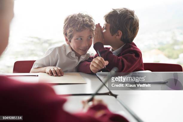 boy whispers in friends ear in classroom at school - mystery fotografías e imágenes de stock