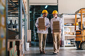Picture of two male warehouse workers with helmets on their heads carrying boxes in their hands. Talking and walking.