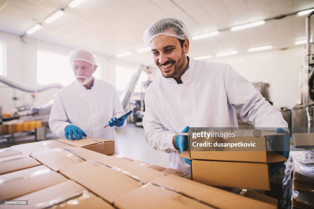 Two male colleagues i sterile clothes preparing boxes with products for transport. Standing in bright room or warehouse and counting package.