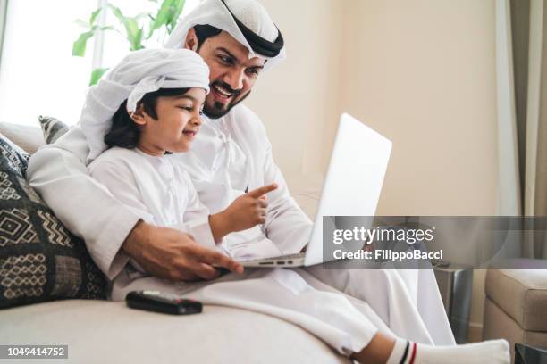 arabische mann mit einem laptop mit seinem sohn auf dem sofa zu hause - emirati guy using laptop stock-fotos und bilder