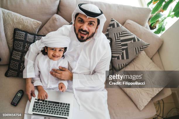 arab man using a laptop with his son on the sofa at home - dish dash stock pictures, royalty-free photos & images