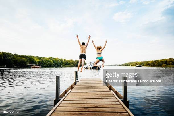 two friends leaping off pier together - lake stock-fotos und bilder