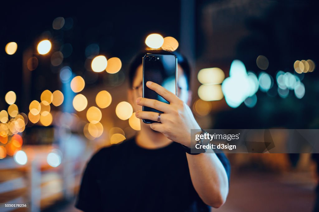 Smart young Asian man taking pictures with smartphone in city street, against illuminated city street light and city traffic