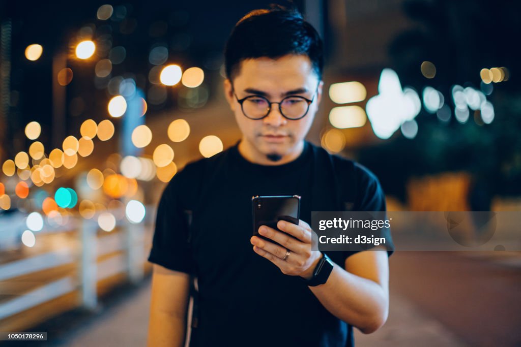 Smart young man text messaging on mobile phone while walking along in city street at night