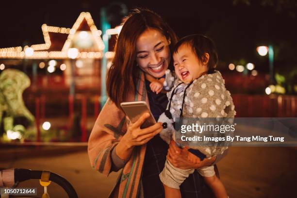 pretty young mom holding baby using smartphone in the park at night joyfully. - mother and daughter on night street stockfoto's en -beelden