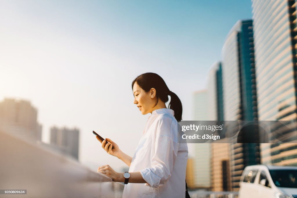 Beautiful young Asian woman texting on smartphone at outdoor parking lot in downtown city