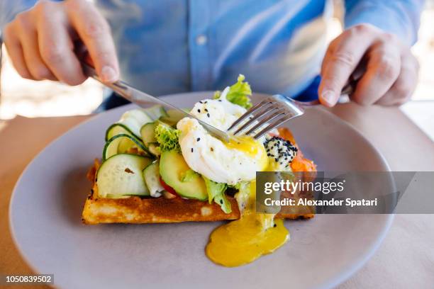 man eating brunch with waffle, avocado, salmon and poached egg with liquid egg yolk - cutting avocado stockfoto's en -beelden