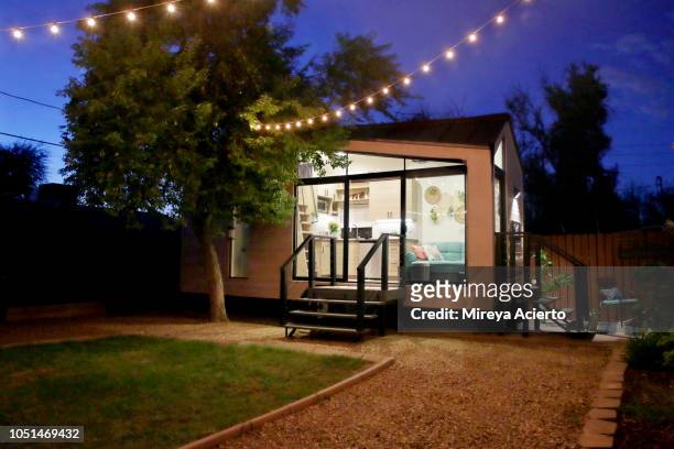a tiny house with large glass windows, sits in the backyard at night, surrounded by trees and party lights. - piccolo foto e immagini stock