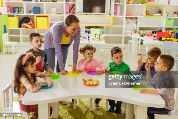 happy teacher with group of children on a lunch break at preschool. - school meal stock pictures, royalty-free photos & images