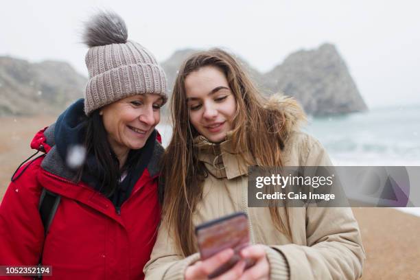 mother and daughter in warm clothing using smart phone on snowy winter beach - winter woman showing stock pictures, royalty-free photos & images