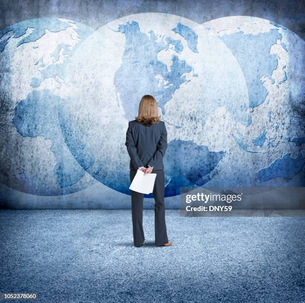 businesswoman looks up at three interlocking globes on wall - political uncertainty stock pictures, royalty-free photos & images