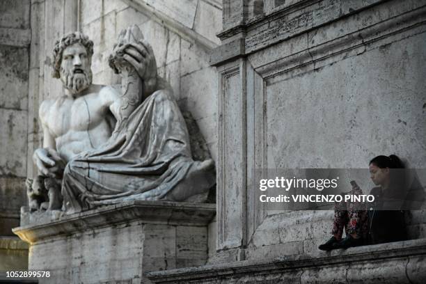 Woman checks her mobile phone while sitting next to a statue representating the river Tiber on Capitol Hill, or Campidoglio, on October 23, 2018 in...