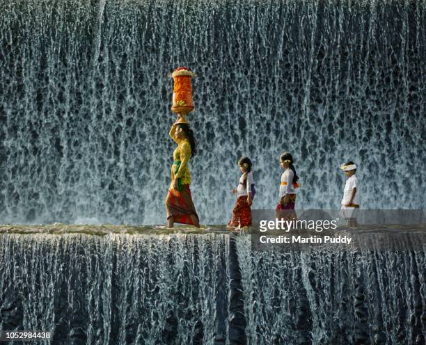 young balinese woman and children wearing traditional clothing, walking across waterfall - bali fotografías e imágenes de stock