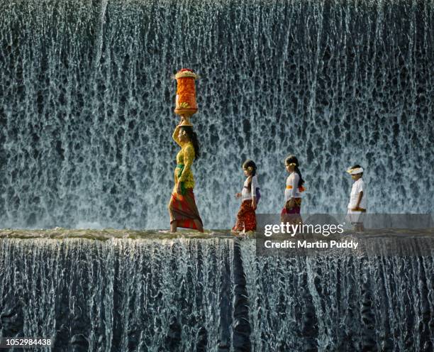 young balinese woman and children wearing traditional clothing, walking across waterfall - portare sulla testa foto e immagini stock