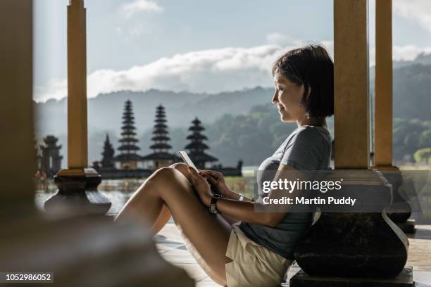 young asian woman sitting inside pavillion using smartphone, in front of tamblingan water temple - indonesia women stock-fotos und bilder