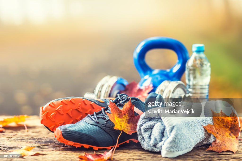 Pair of blue sport shoes water and  dumbbells laid on a wooden board in a tree autumn alley with maple leaves -  accessories for run exercise or workout activity.