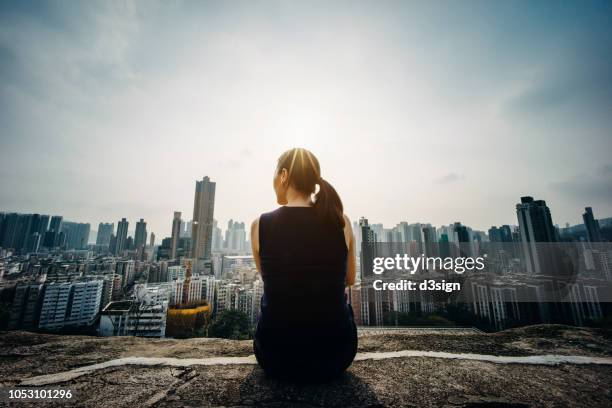 young woman sitting on the top of mountain enjoying the panoramic view of spectacular city skyline - ergens overheen kijken stockfoto's en -beelden