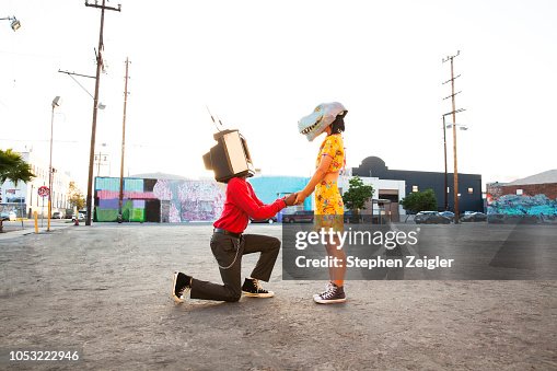 Man with an old TV set over his head proposing to a woman wearing a dinosaur mask Man with an old TV set over his head proposing to a woman wearing a dinosaur mask