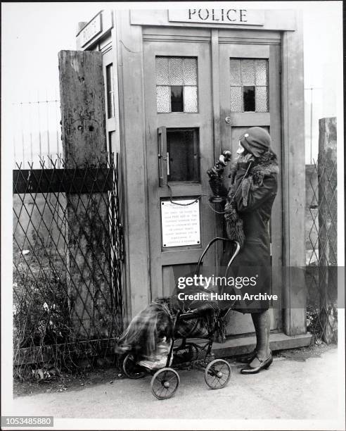 Woman with a baby in a pushchair uses the telephone in an Emergency Police Call Box, London, England, 1930s. FOX F 38133.