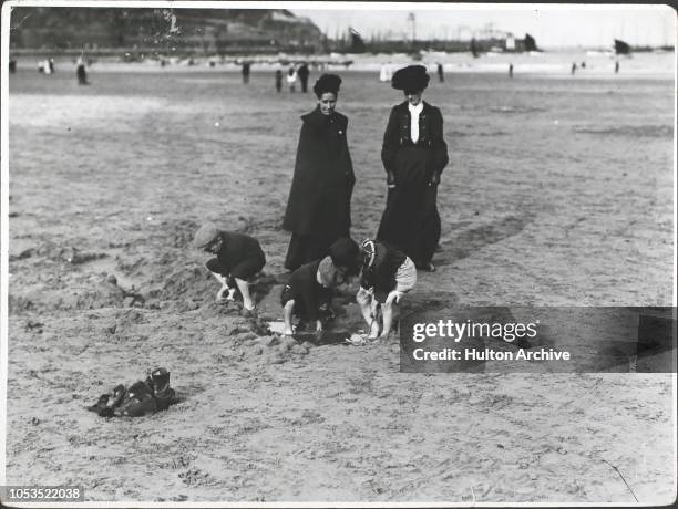 Two women watching children at play, digging on the beach on Scarborough sands, England, 1907.