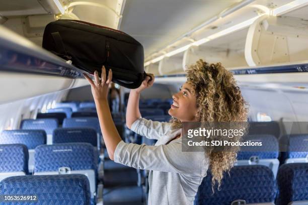 young woman places luggage in airline overhead bin - hand luggage stock pictures, royalty-free photos & images