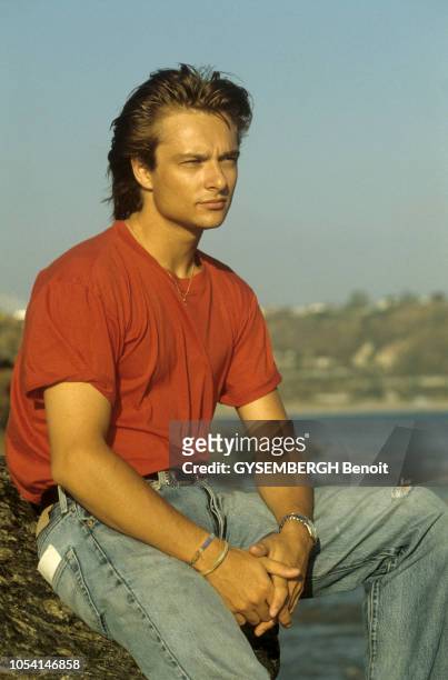 Californie, Etats-Unis - Août 1988 - David HALLYDAY, en tee-shirt rouge et jeans déchiré, posant de trois-quarts, assis sur un rocher.