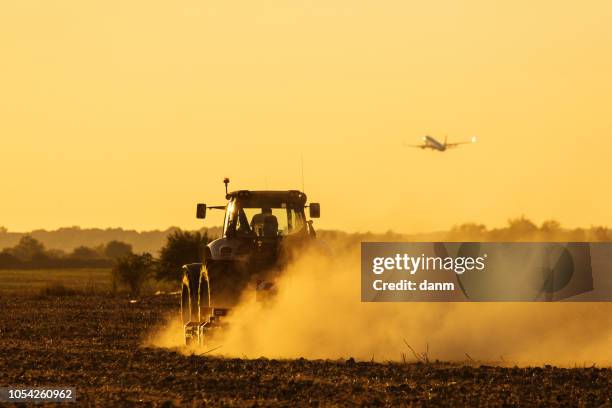 modern tractor plowing at the sunset with lot of dust in background and with a plane taking off - plough stock pictures, royalty-free photos & images