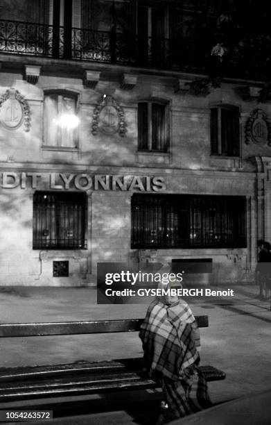 Paris, France, circa 1985 --- Un sans domicile fixe âgé, emmitouflé dans une couverture, passant la nuit dans la rue, assis sur un banc public situé...