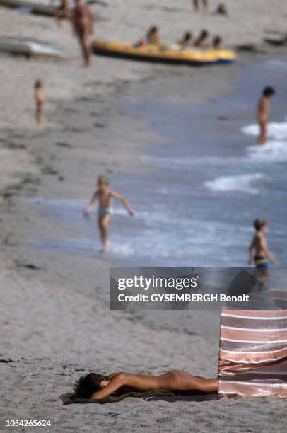 Bretagne, août 1983 - Nudistes sur les plages du littoral breton. Femme nue bronzant, à plat ventre. Des enfants jouent dans l'eau à l'arrière plan.