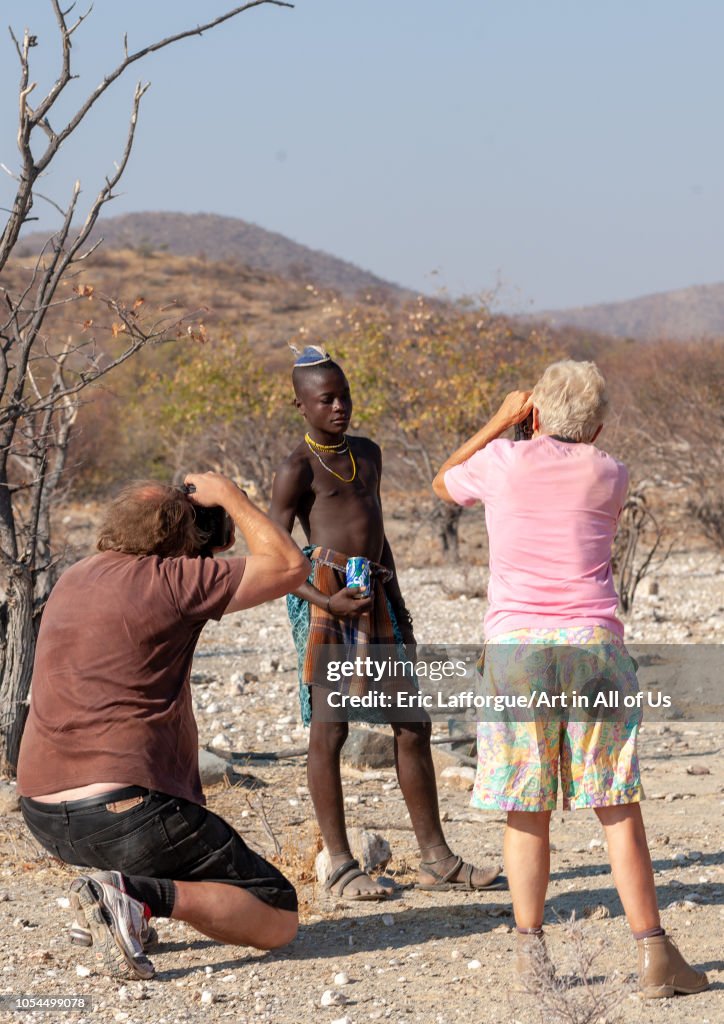 Western tourists taking pictures of a Himba tribe young man, Cunene Province, Oncocua, Angola...