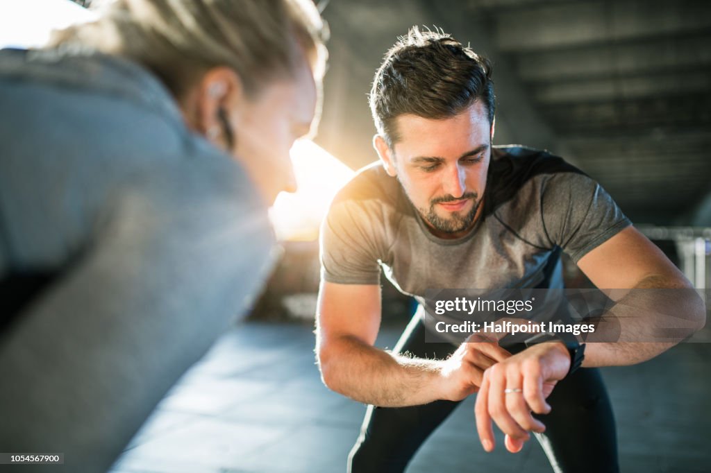 Young sporty man and woman runners resting after the run under the bridge outside in a city.