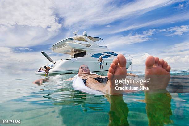 man floating on air bed with boat in background - jacht stock-fotos und bilder