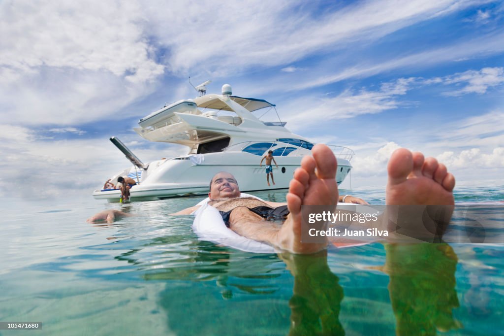 Man floating on air bed with boat in background